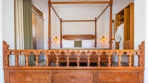 A four-poster bed with a wooden frame and intricate carvings, flanked by a wooden bench with drawers.