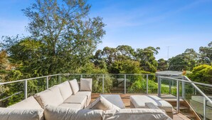 A sunlit outdoor seating area with a sofa and cushions, surrounded by a glass railing and overlooking a lush landscape.