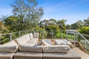 A sunlit outdoor seating area with a sofa and cushions, surrounded by a glass railing and overlooking a lush landscape.