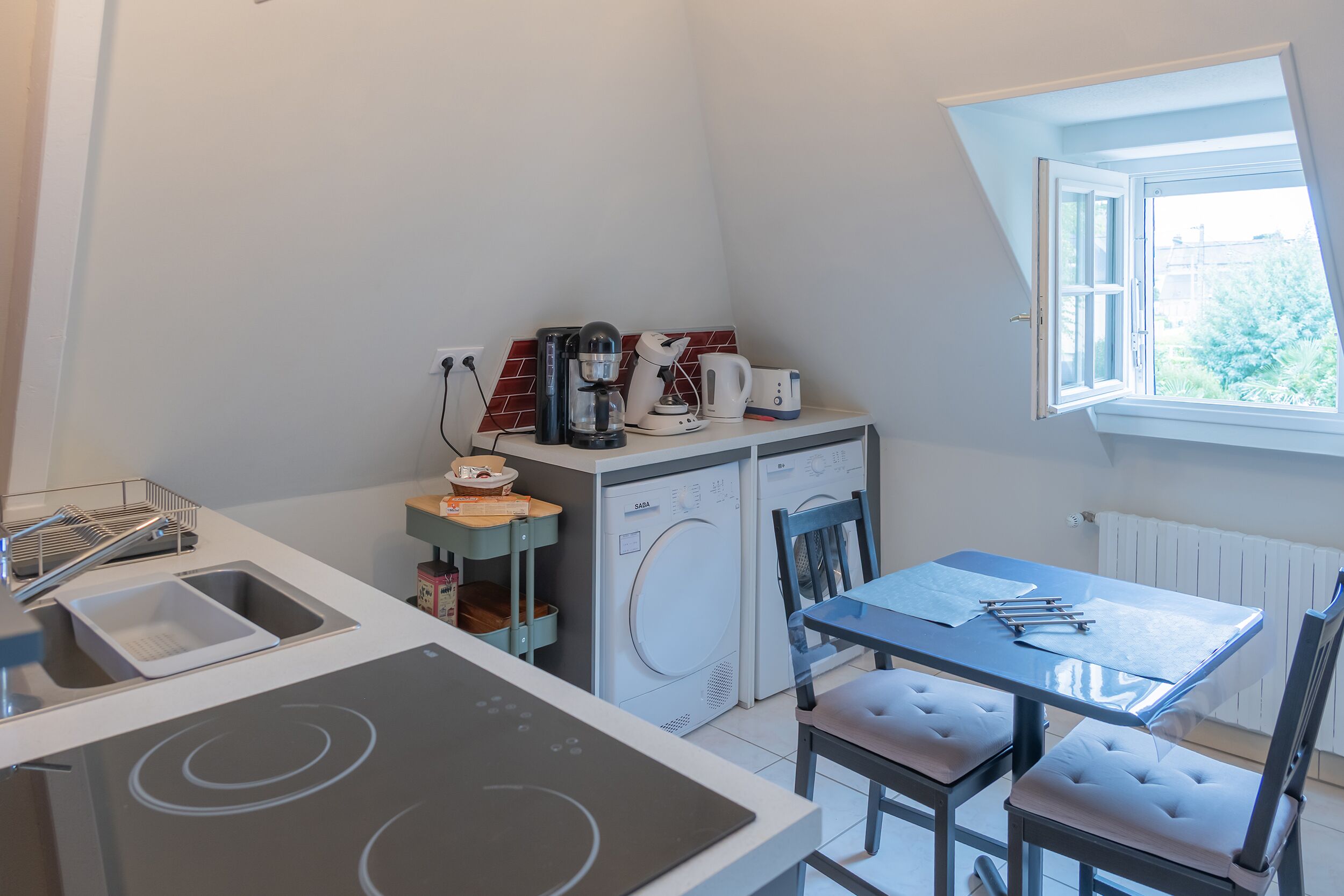 Eine kompakte Küche mit Einbauofen, Kaffeemaschine, Wasserkocher und Waschmaschine. Ein kleiner Tisch mit Stühlen und ein Fenster sorgen für natürliches Licht.