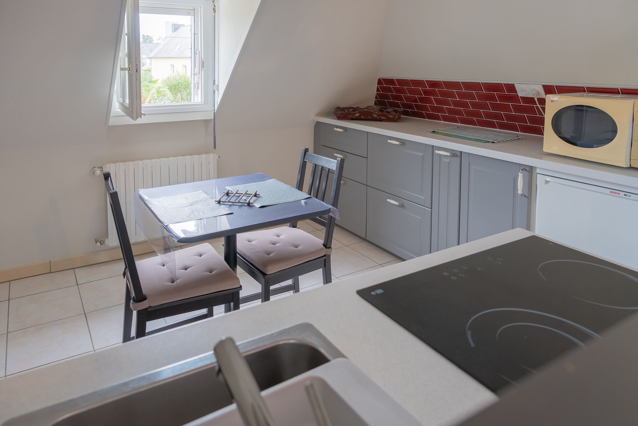 A compact kitchen with a dining table, chairs, a Bosch microwave, and a red-tiled backsplash.