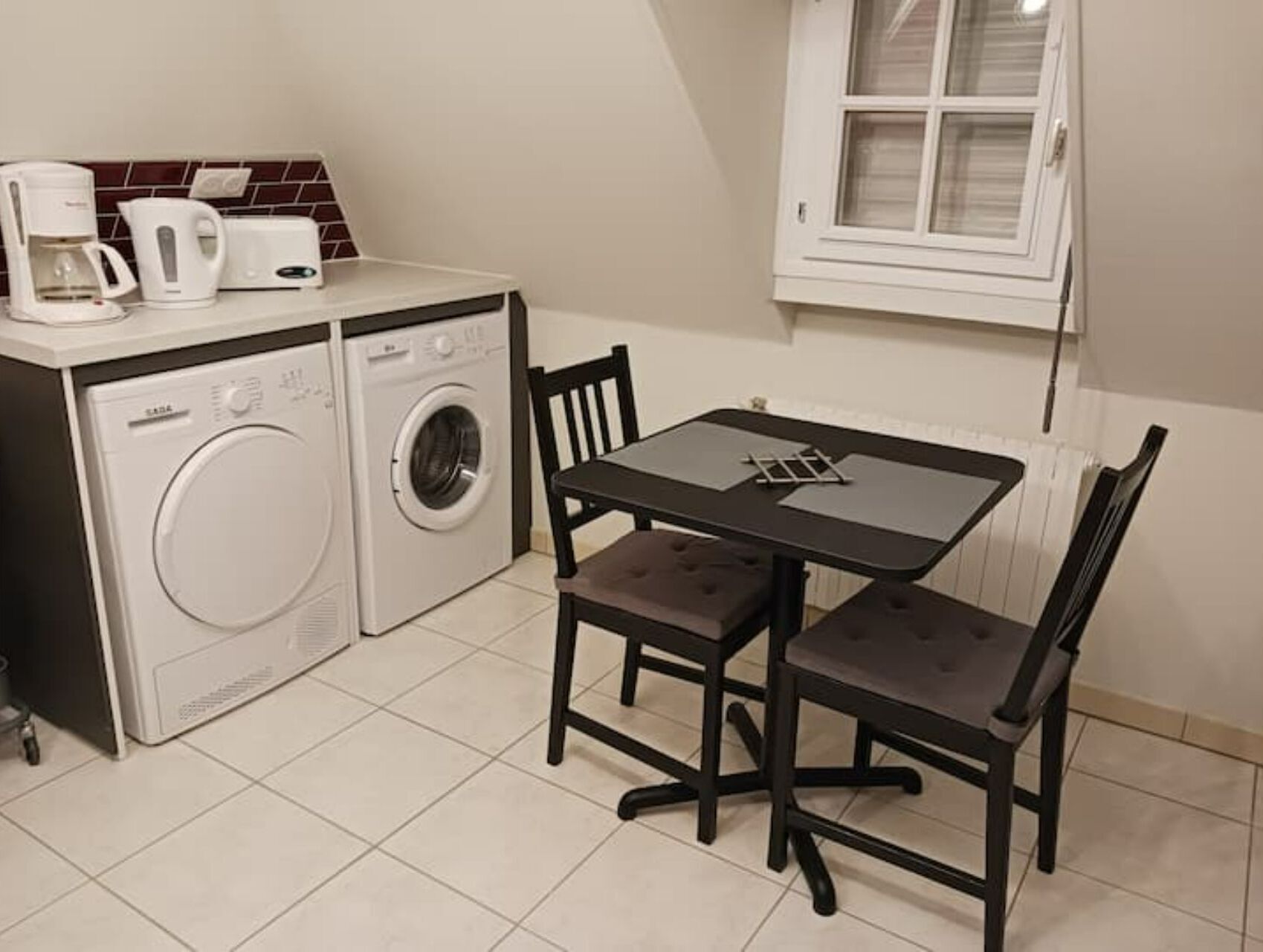 Une cuisine compacte équipée d’un lave-linge, d’une machine à café et d’un grille-pain. On y trouve une petite table avec deux chaises et une fenêtre au-dessus.