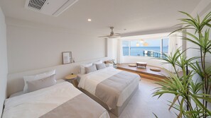 Una habitación de hotel moderna con dos camas, un ventanal con vistas al mar, un ventilador de techo y una mesita de noche con lámpara.