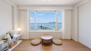 Una habitación con piso de madera, una mesa redonda, dos otomanas tejidas, una lámpara blanca y vistas al mar a través de un amplio ventanal con cortinas.