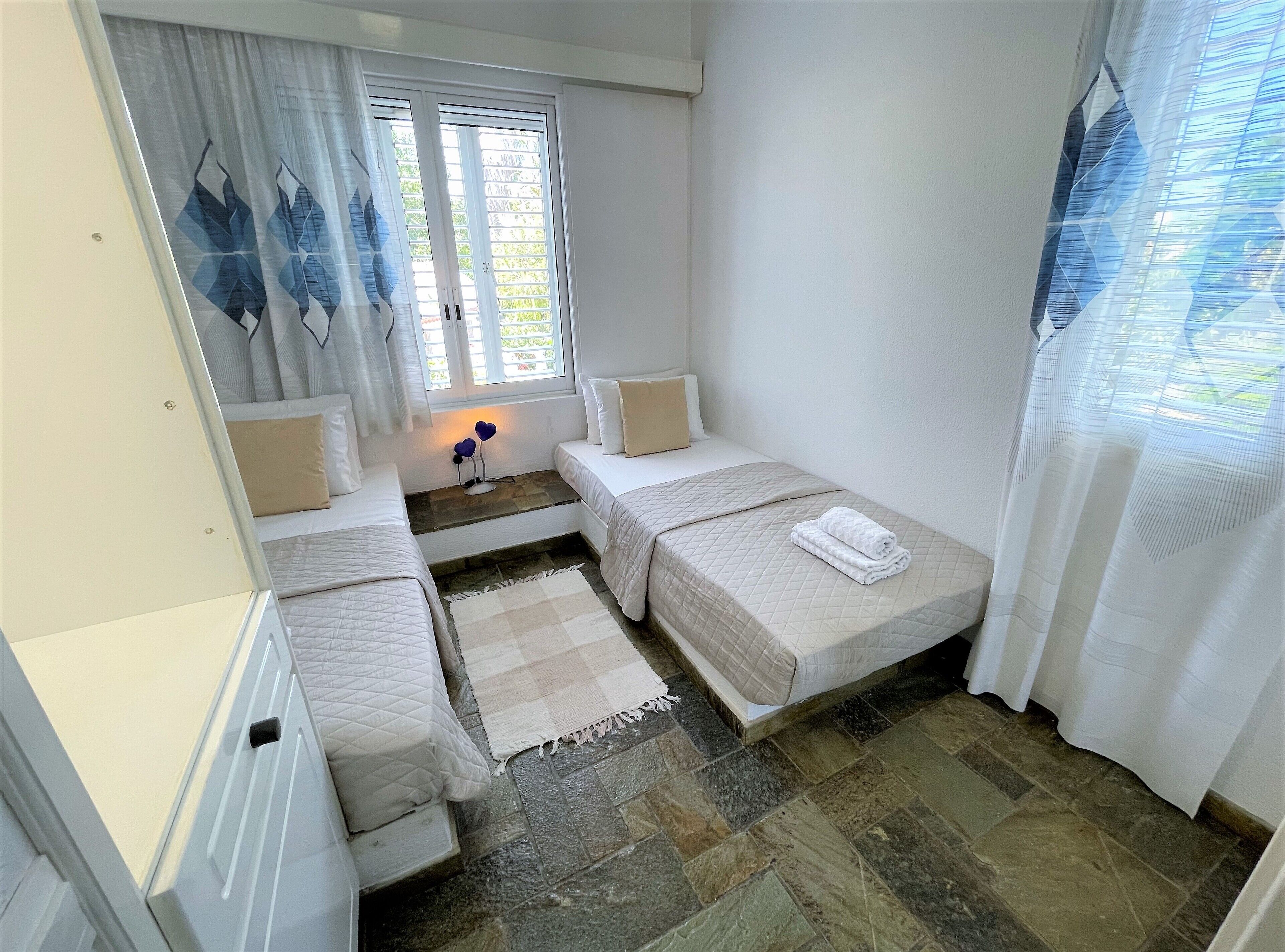 A small, cozy room with two single beds, a window with curtains, a stone floor, and a small table with a lamp.