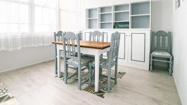 Un comedor con mesa de madera y sillas grises, un armario blanco con estantes, y una alfombra estampada.