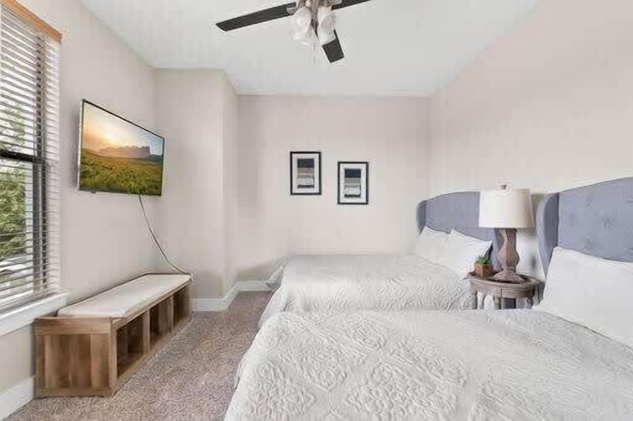 Een slaapkamer met twee bedden, een plafondventilator, een bankje, een televisie en ingelijste foto's aan de muur.