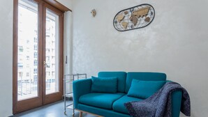 Un canapé de couleur turquoise, recouvert d’une couverture, une décoration représentant une carte du monde et une vue sur des bâtiments à travers une porte.