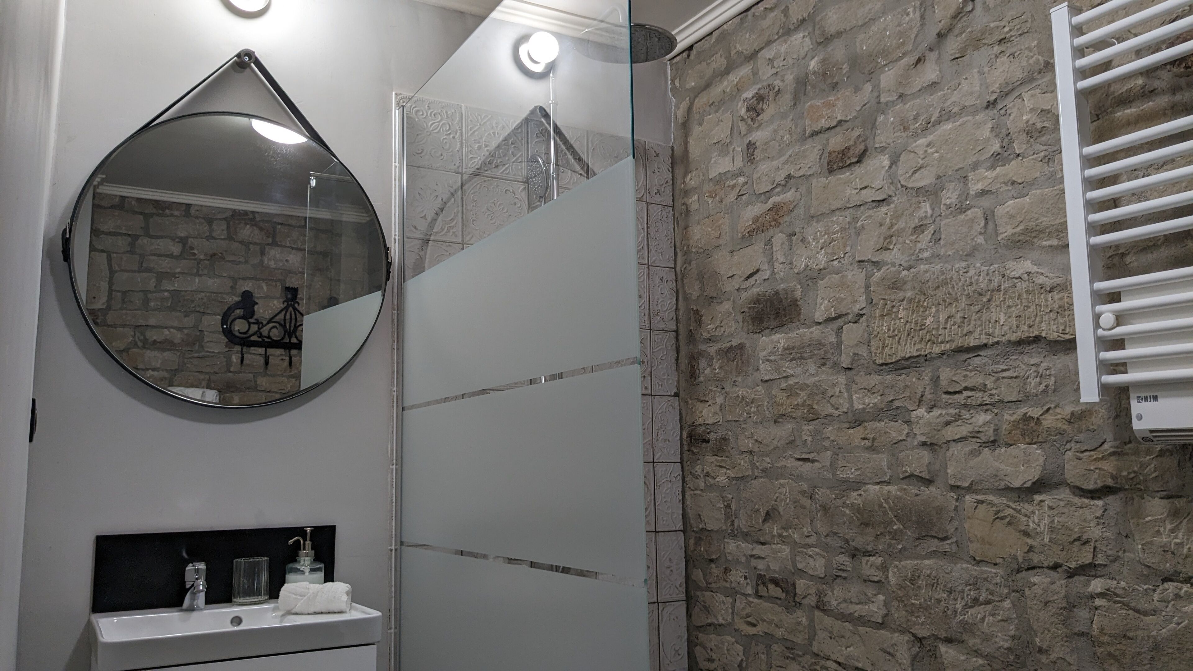 Un baño con pared de piedra, un espejo redondo, cabina de ducha de vidrio y un perchero blanco.