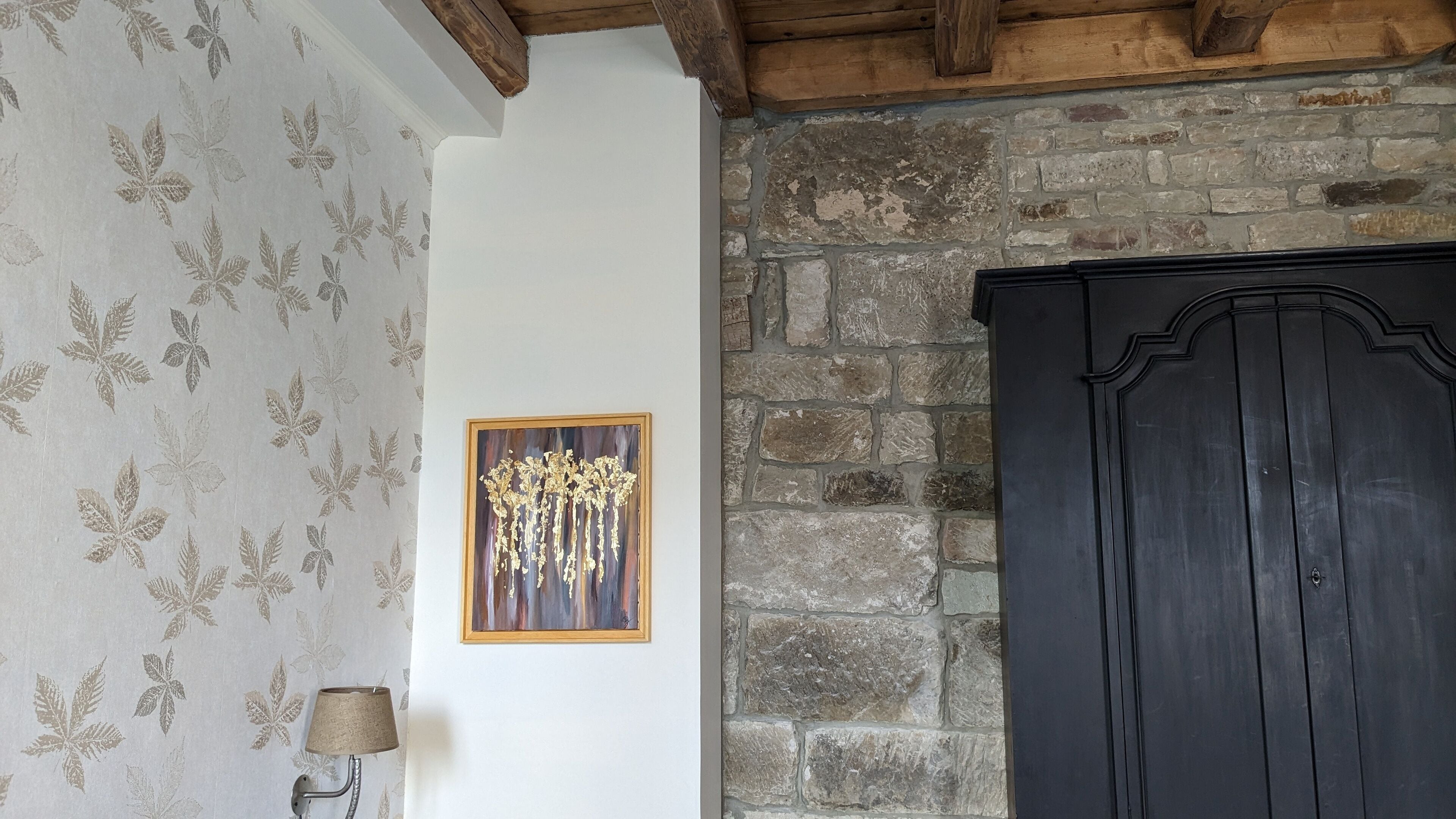 Una habitación con papel tapiz estampado, una obra de arte enmarcada, una lámpara y un armario de madera oscura frente a una pared de piedra.