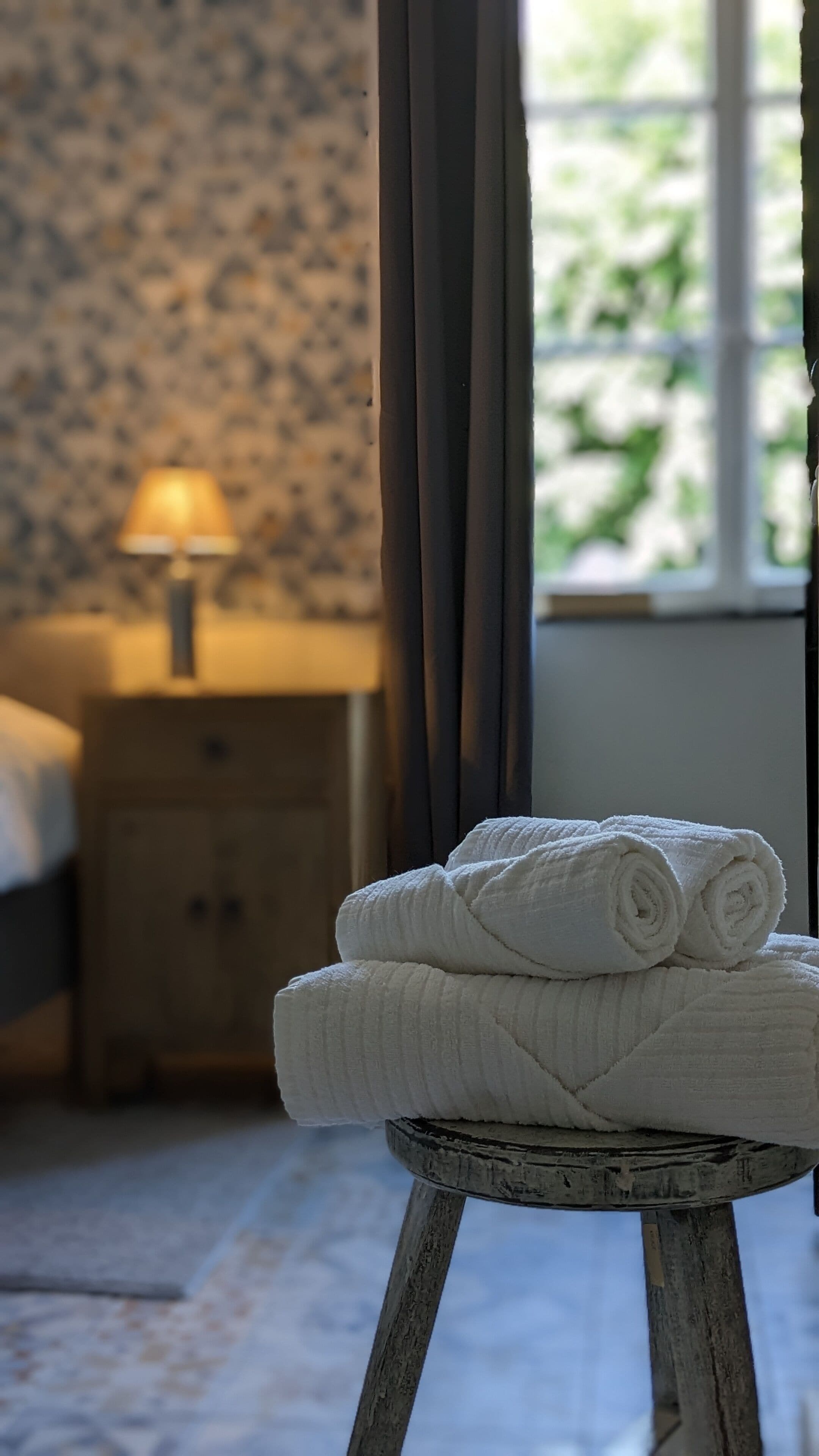 Een net opgemaakt bed met een lamp op een nachtkastje, een raam met gordijnen en een stapel handdoeken op een kruk.