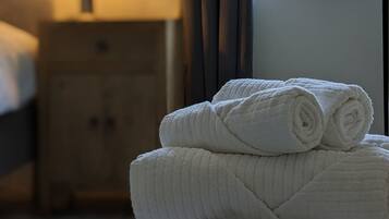 Een net opgemaakt bed met een lamp op een nachtkastje, een raam met gordijnen en een stapel handdoeken op een kruk.