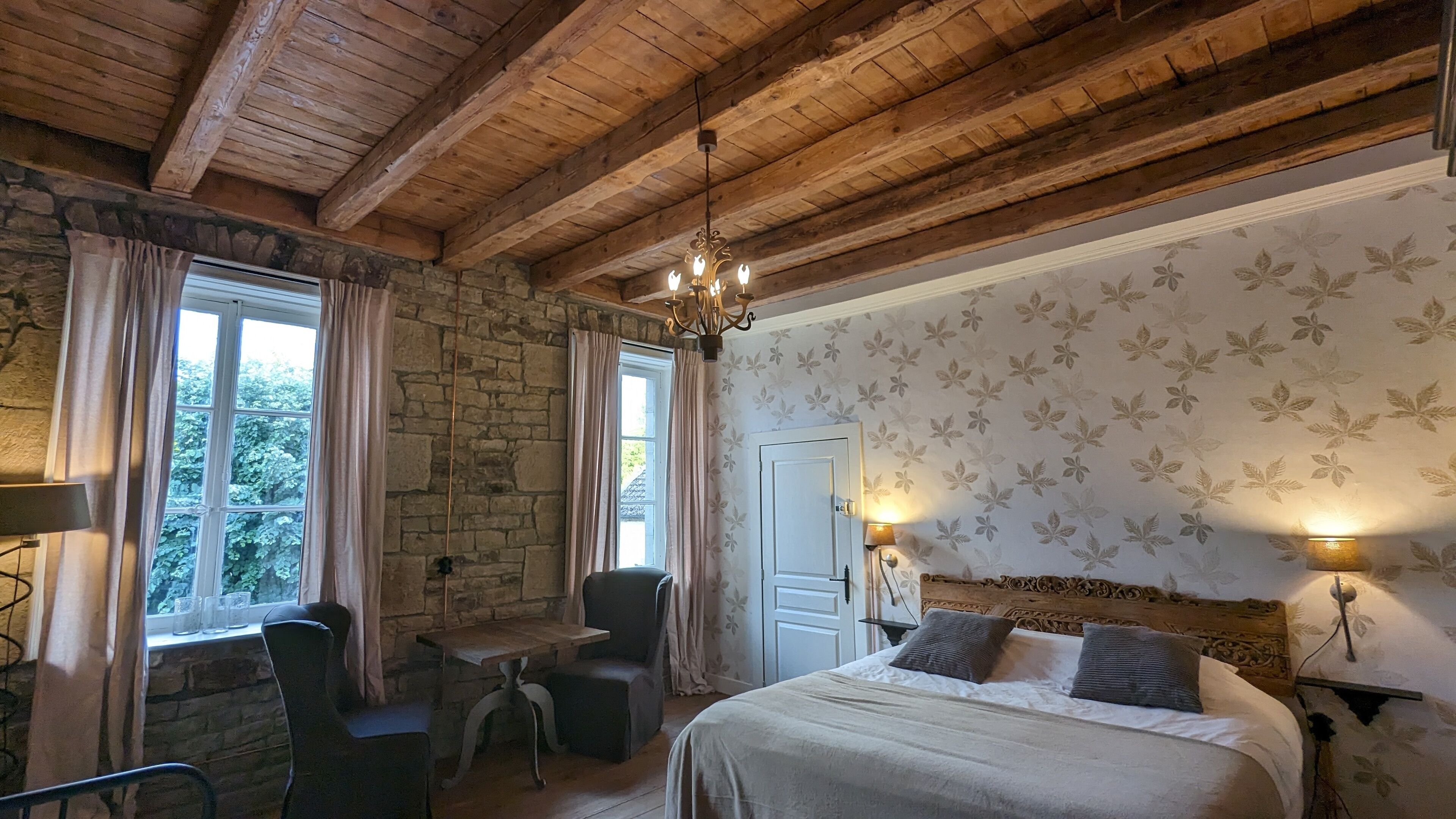 Een kamer met een stenen muur, houten balken in het plafond, een bed met kussens, een tafel met stoelen en een raam met gordijnen.