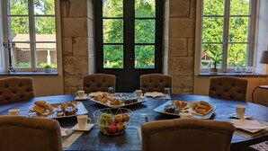 Una mesa de comedor lista para el desayuno, con pasteles, fruta y tazas de café, rodeada por cuatro sillas tapizadas, con vistas a la vegetación a través de amplias ventanas.