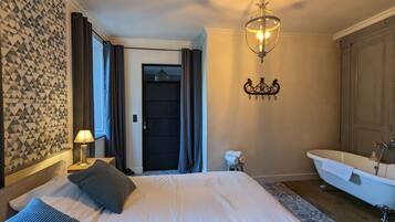 Un dormitorio con cama, lámpara de noche, candelabro, baño con bañera y puerta que da a otra habitación.