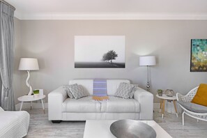 A modern living room with a white sofa, two side tables, a coffee table, and a painting of a tree on the wall. - Witsand 804 (Cape Town)