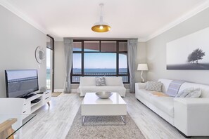 Uma sala de estar moderna com uma TV de tela plana, um sofá branco, uma mesa de centro branca e uma grande janela com vista para o mar.