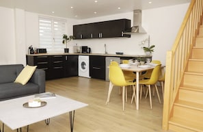 A modern kitchen with dark cabinets, a washing machine, and a dining area with yellow chairs and a white table.
