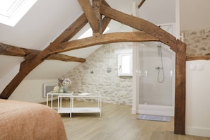 A room with a sloped ceiling, exposed wooden beams, a stone wall, a shower, a white bed, and a small table with flowers.