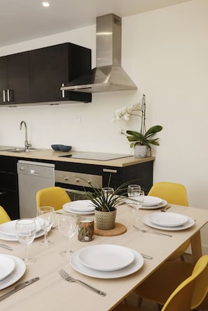 A modern kitchen with dark cabinets, a stainless steel range hood, a dishwasher, and a dining table set with plates, glasses, and a plant.