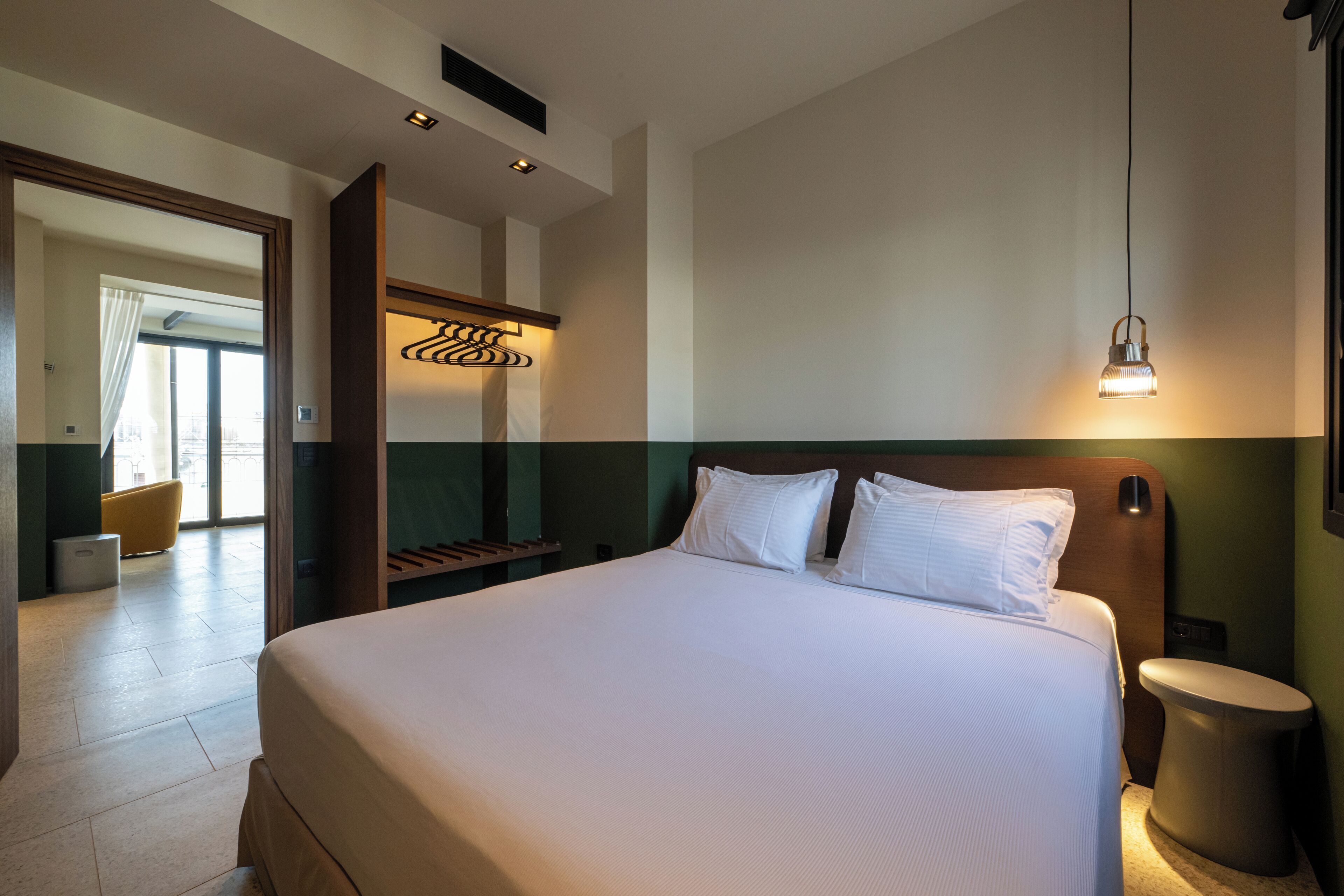 A hotel room with a large bed, a bedside table, a wall-mounted light fixture, and a view of the outside through a window.