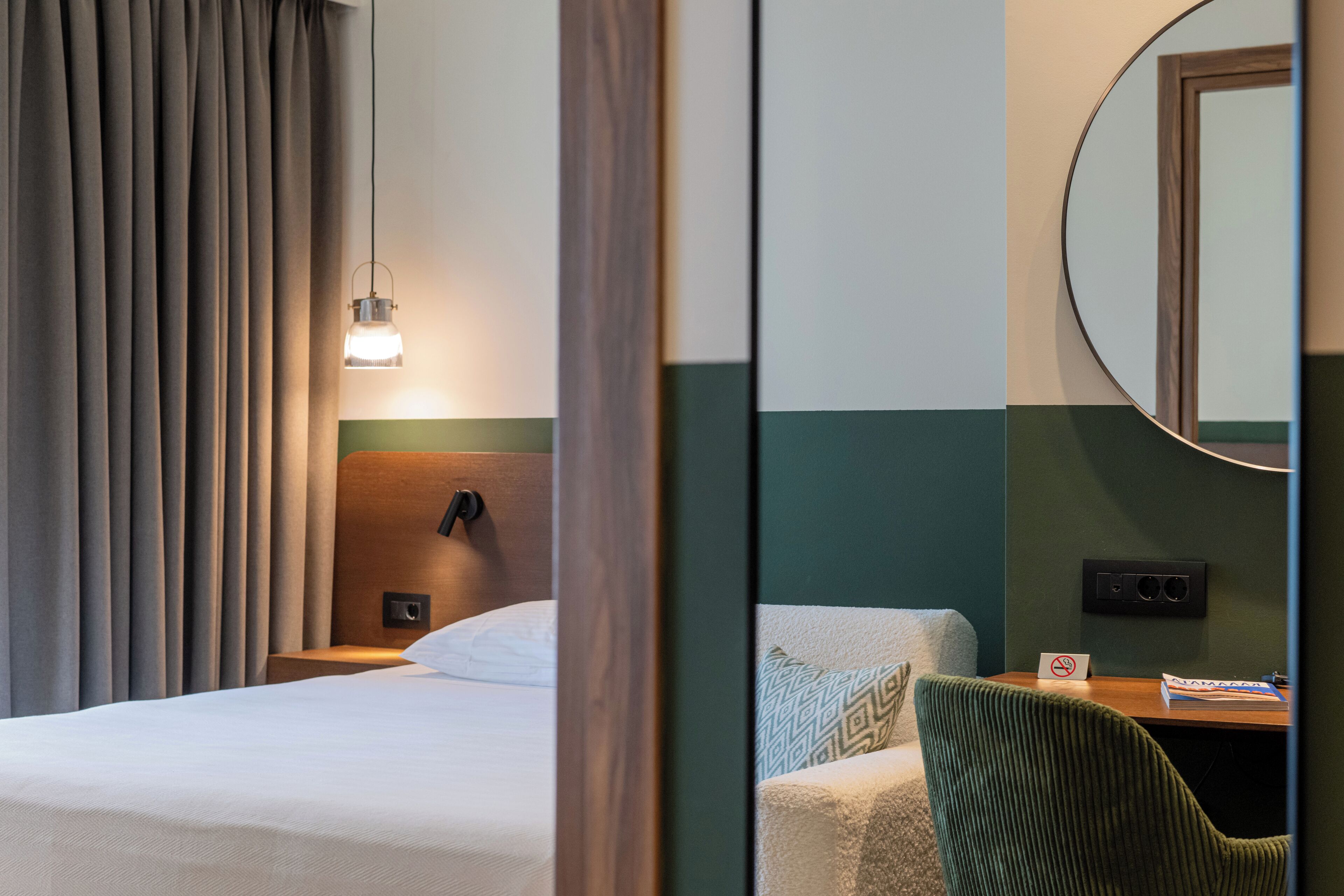 Une chambre d’hôtel comprenant un lit, une table de chevet, un bureau avec une chaise, un miroir et une fenêtre avec des rideaux.