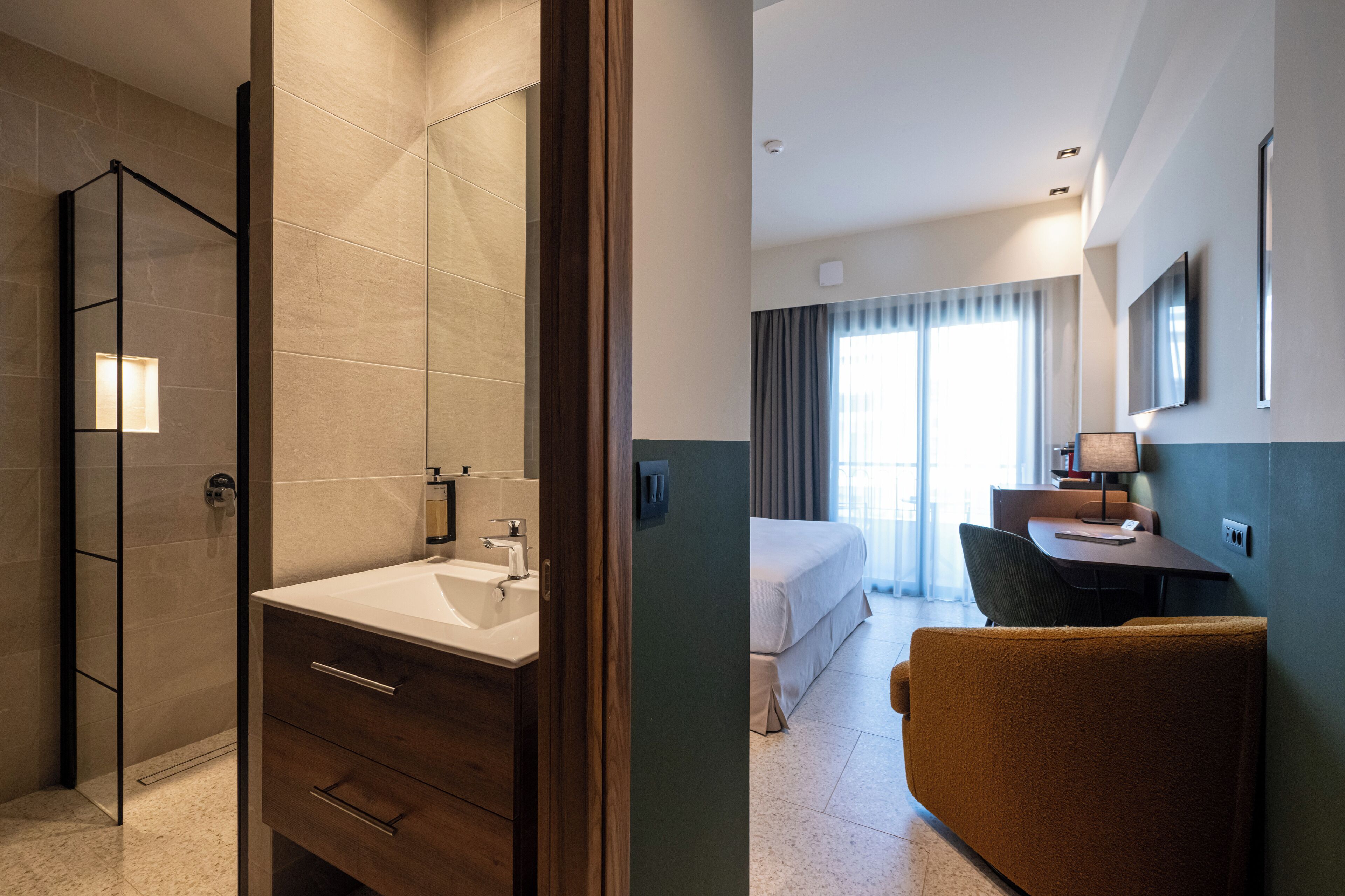Hotelkamer met een badkamer, voorzien van een glazen douchecabine, een houten wastafelmeubel met een wastafel, en een bed met uitzicht op het landschap.