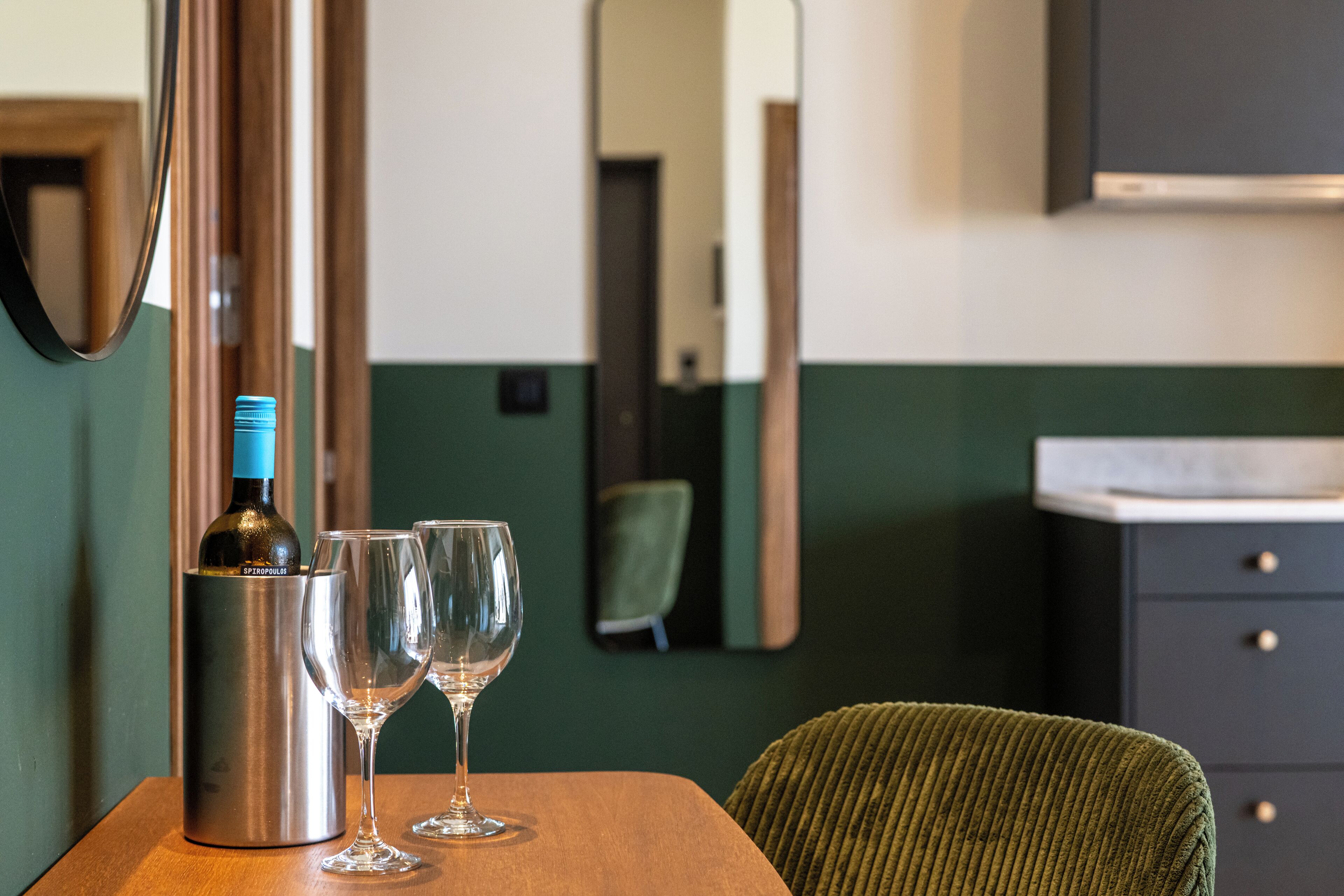 Una habitación con una mesa de madera, una botella de vino y dos copas. Se ve una silla verde y un armario oscuro al fondo.