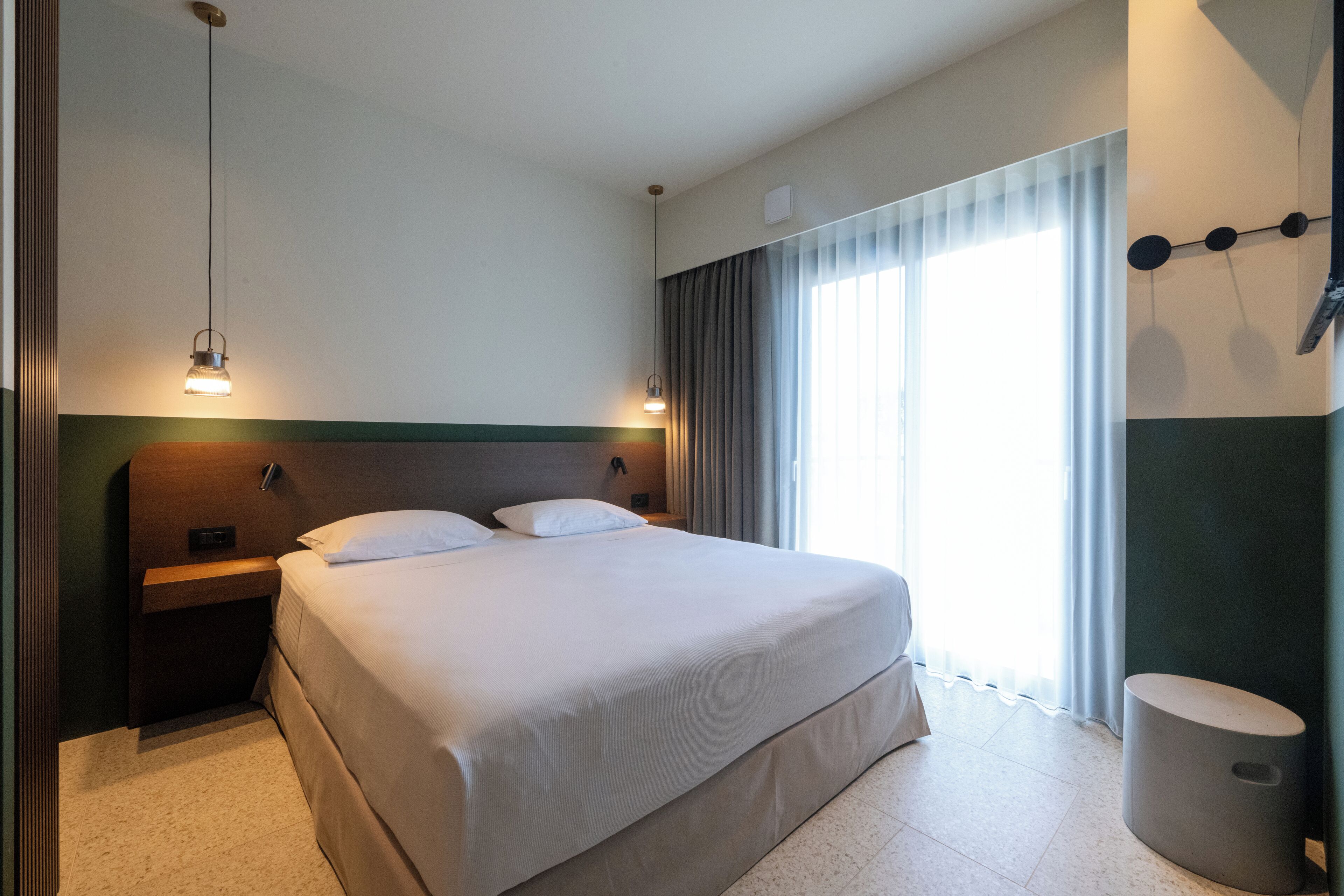A hotel room with a large bed, bedside tables, a nightstand, a lamp, a window with curtains, and a wall-mounted light fixture.