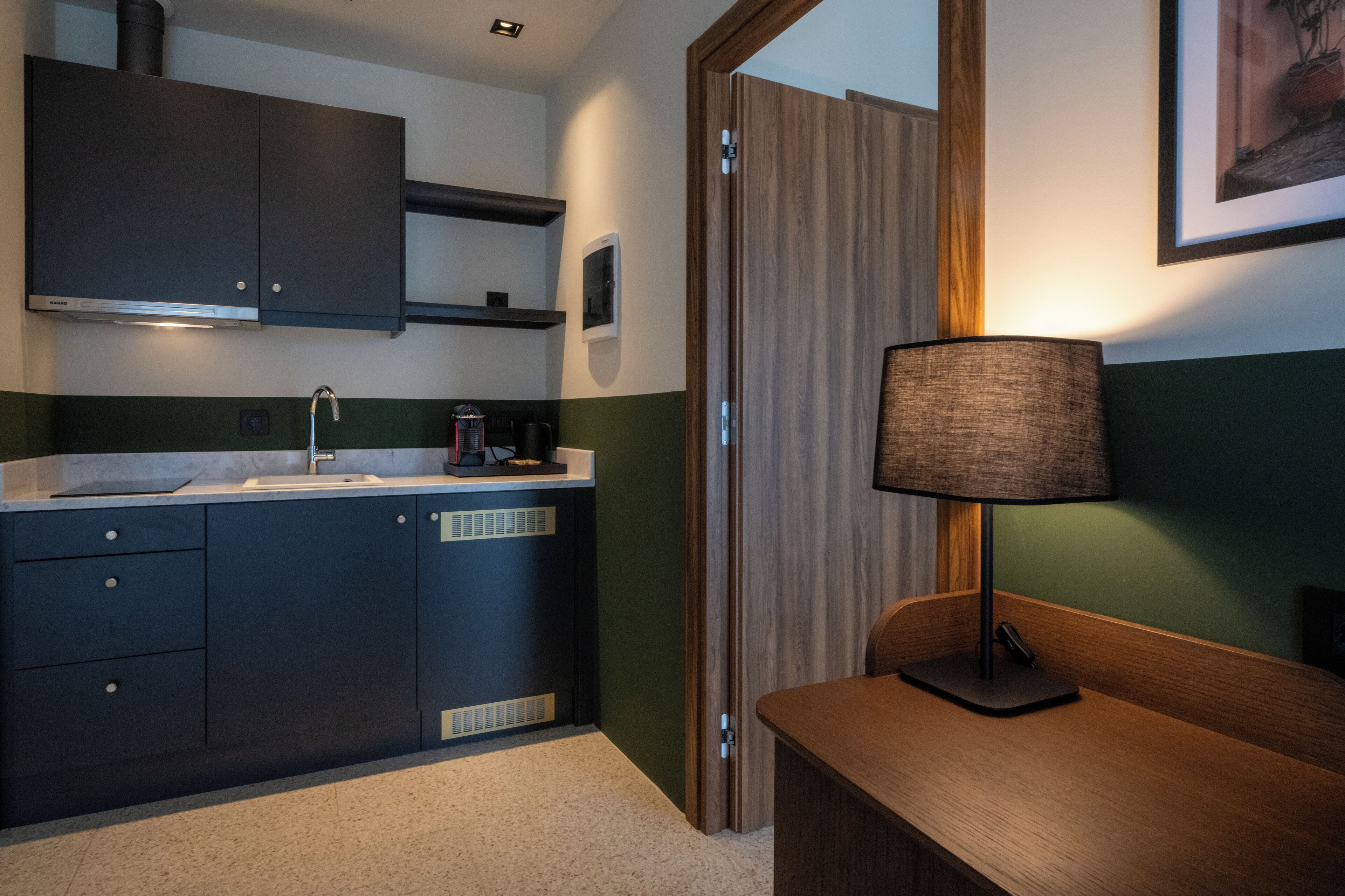 A modern kitchen with dark cabinets, a sink, and a countertop. There is a wooden door and a lamp on a wooden table.
