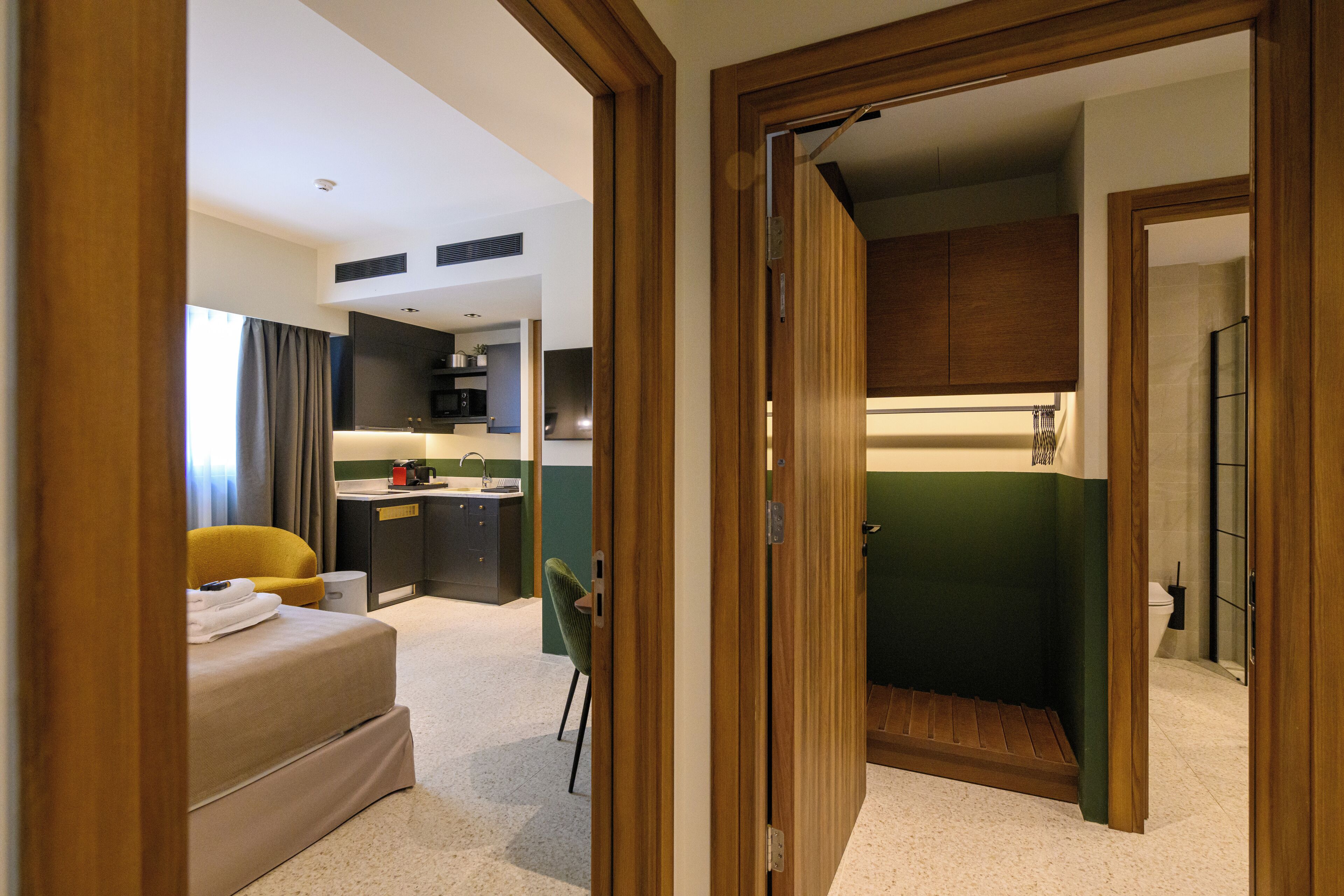 A modern hotel room with a bed, a small desk, a chair, a kitchenette, and a bathroom.