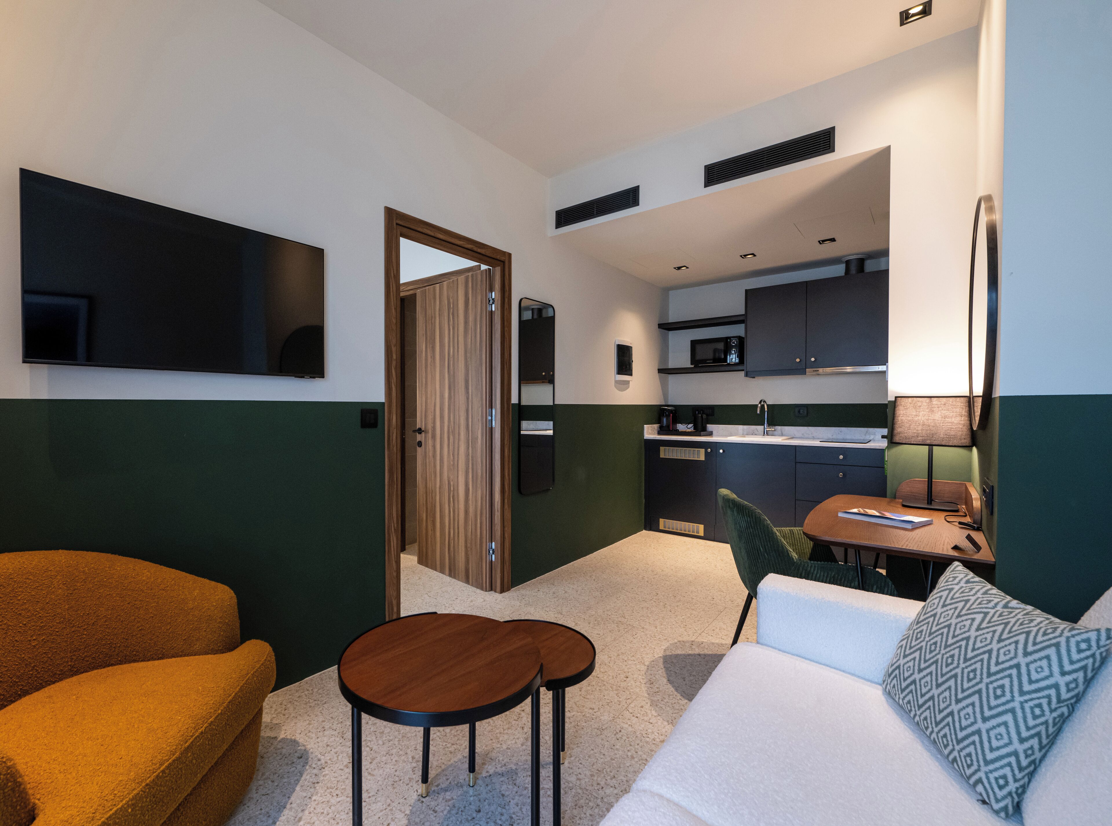 Un espacio habitable compacto con un sofá, una mesita, un televisor, una kitchenette y una puerta que da a otra habitación.
