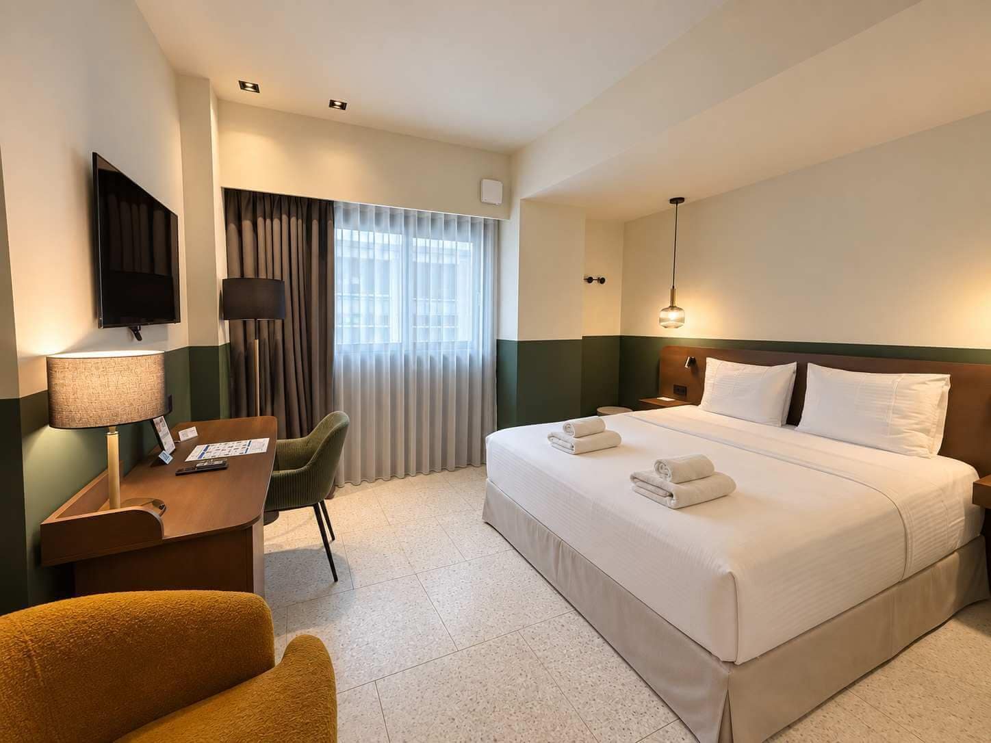 Quarto de hotel com cama, escrivaninha, cadeira, televisão e janela com cortinas.