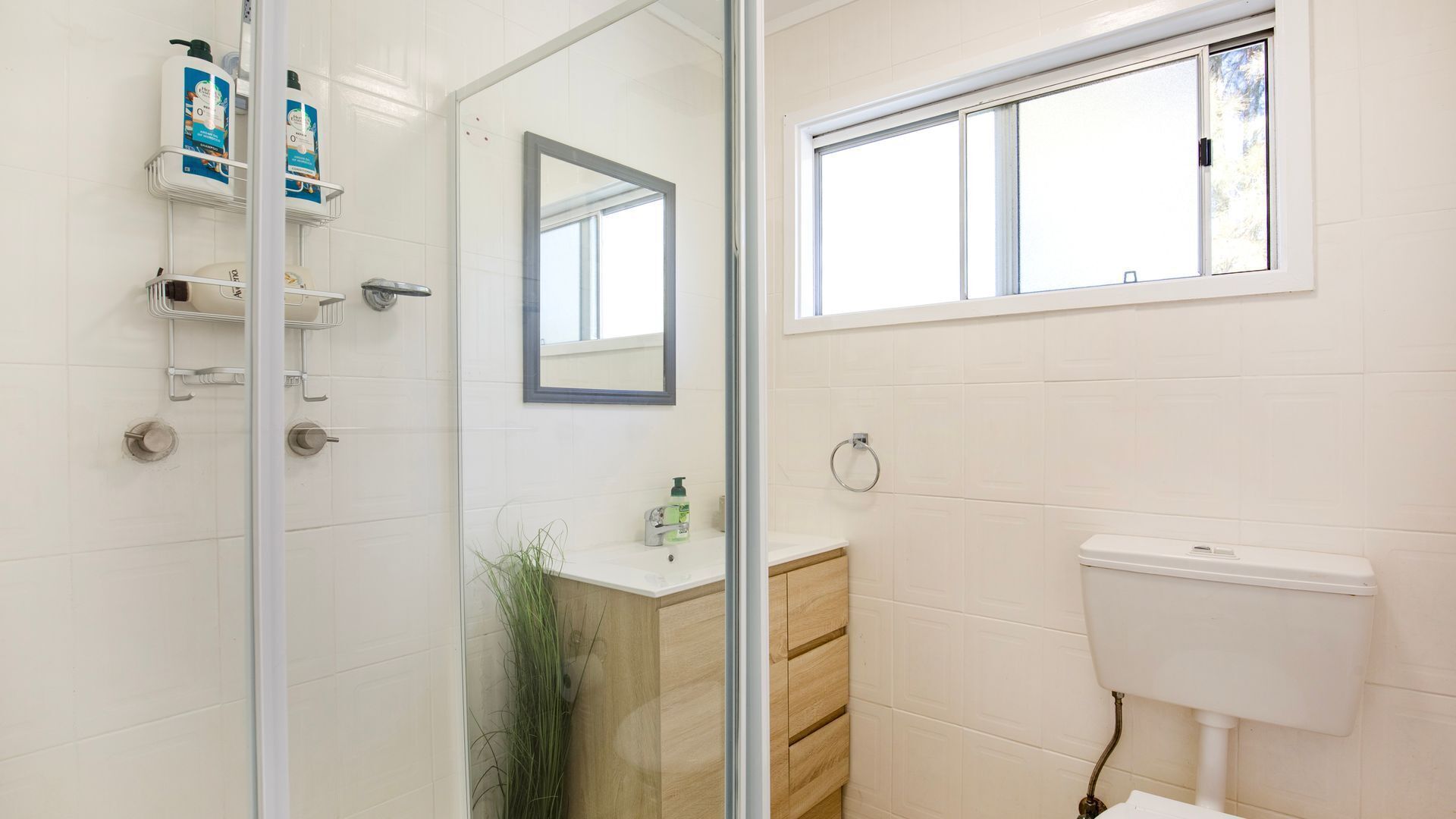 A bathroom with a glass shower door, a toilet, a sink with a mirror, and toiletries on a shelf.