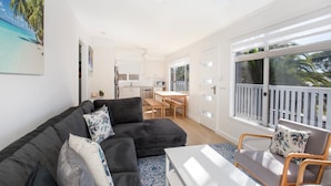 A modern living room with a dark grey sofa, a white coffee table, and a wooden chair. There is a kitchen area with white cabinets and a dining table. Large windows provide a view of the outdoors.