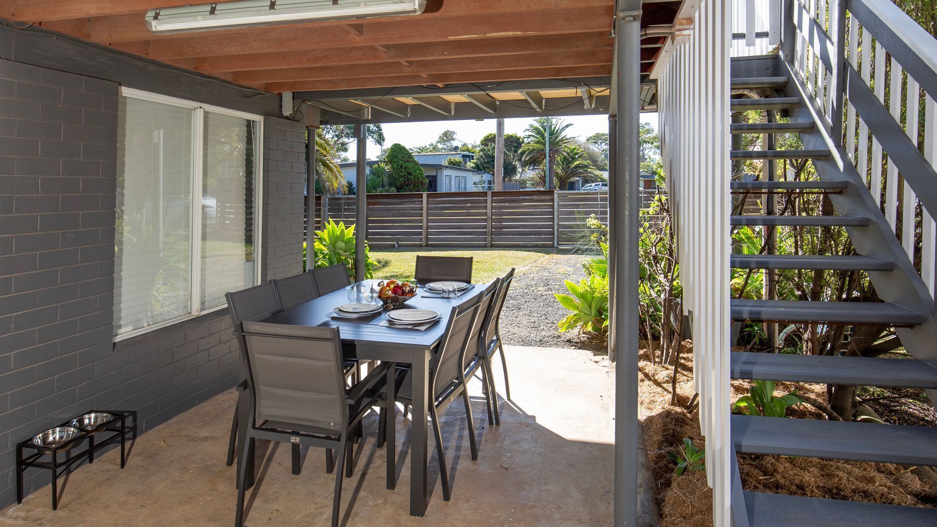Un patio exterior con una mesa lista para comer, rodeado de sillas y una escalera que lleva a un nivel superior.