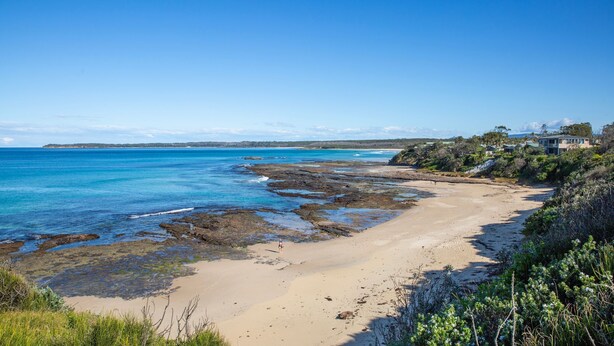 A coastal beach with rocky areas, clear blue water, and houses on the right.