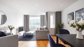 A modern living room with a sofa, a coffee table, and a dining table with chairs. There is a large window with curtains, a flat-screen TV, and a mirror on the wall. - PBC | One Bedroom Apartment with Balcony JT4 (London)