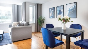 A modern living room with a grey sofa, blue chairs, a dining table, and abstract art on the wall. - PBC | One Bedroom Apartment with Balcony JT4 (London)
