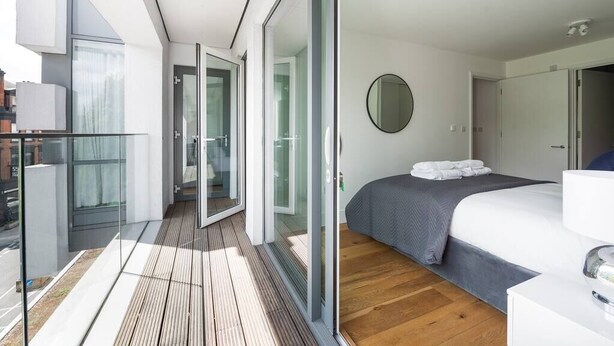 A modern hotel room with a balcony, a bed with a grey bedspread, and a round mirror on the wall. - PBC | One Bedroom Apartment with Balcony JT2 (London)