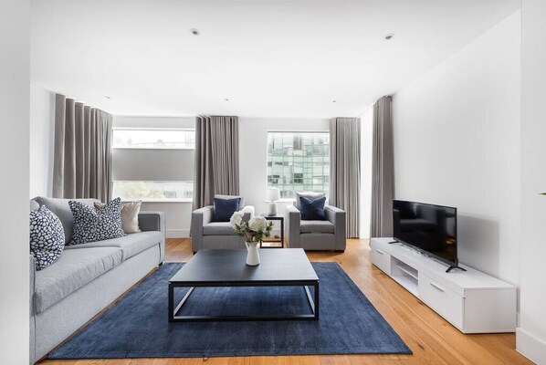 A modern living room with a sofa, two armchairs, a coffee table, a TV, and large windows.