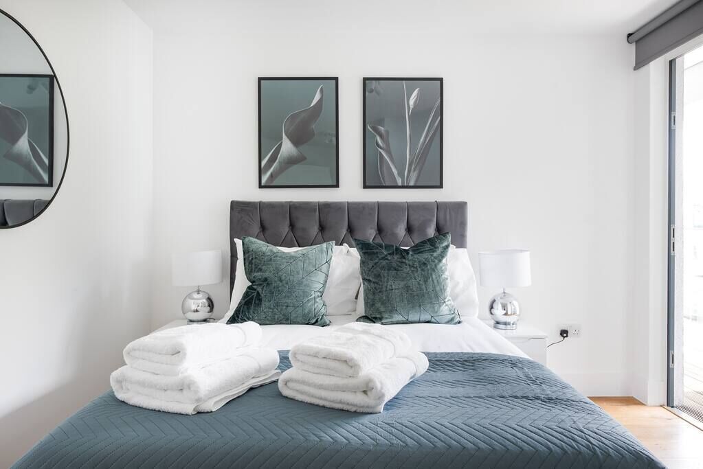 A modern bedroom with a grey upholstered headboard, two framed wall art pieces, a neatly made bed with white towels, and a bedside lamp.
