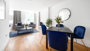 A modern living room with a dining area, featuring a sofa set, a coffee table, and a dining table with chairs. - PBC | One Bedroom Apartment with Balcony JT2 (London)