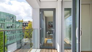 Un appartement moderne doté d’un balcon, d’une porte vitrée donnant sur l’espace de vie et offrant une vue sur un immeuble verdoyant situé de l’autre côté de la rue.