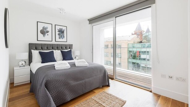 Une chambre moderne avec un grand lit, une table de chevet, une fenêtre donnant sur des immeubles et deux œuvres d’art encadrées accrochées au mur.