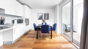 A modern kitchen with white cabinets, a dining table with blue chairs, and a large sliding glass door leading to an outdoor area. - PBC | One Bedroom Apartment with Balcony JT6 (London)
