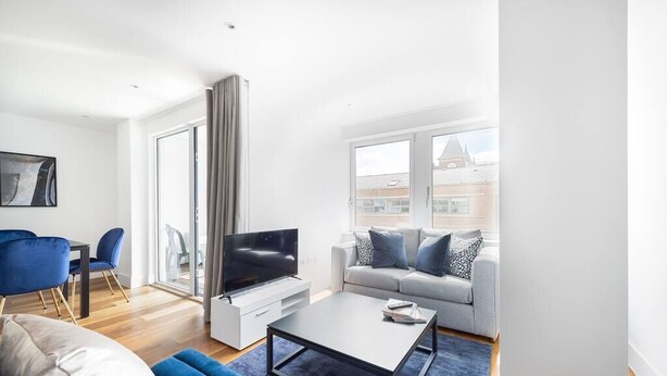 A modern living room with a flat-screen TV, a grey sofa with blue cushions, a coffee table, and a dining area with blue chairs. - PBC | One Bedroom Apartment with Balcony JT6 (London)