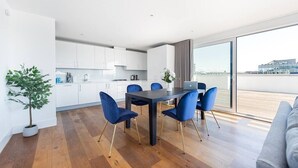 A modern kitchen with white cabinets, a dining table with blue chairs, and a large window offering a view of the city. - PBC | Three Bedroom Apartment with Balcony JT9 (London)