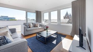 A modern living room with a sofa, coffee table, and TV, large windows with a city view, and a balcony. - PBC | Three Bedroom Apartment with Balcony JT9 (London)