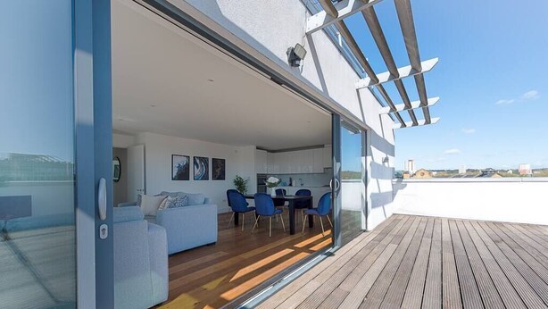 A modern living area with a glass door leading to a balcony, a dining area, and a kitchen visible through the glass. - PBC | Three Bedroom Apartment with Balcony JT9 (London)